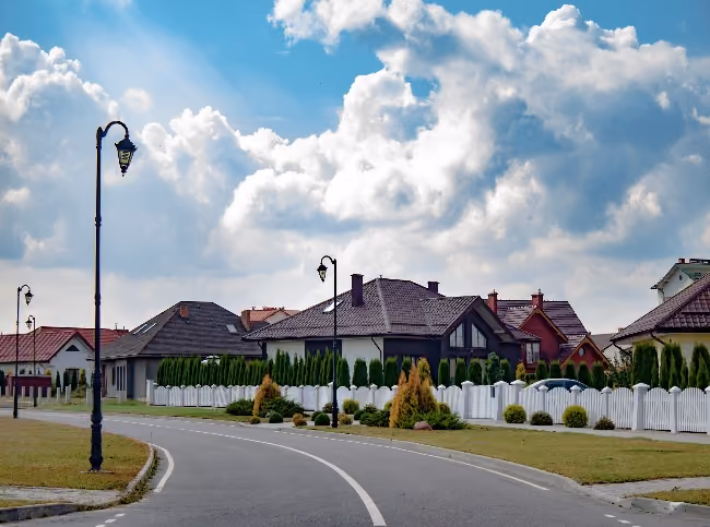 Curved street lined with modern houses, white fences, lampposts, and cloudy blue sky overhead.