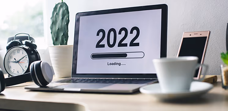 Laptop screen shows 2022 with a loading bar; desk has alarm clock, headphones, cactus, phone, and coffee cup.