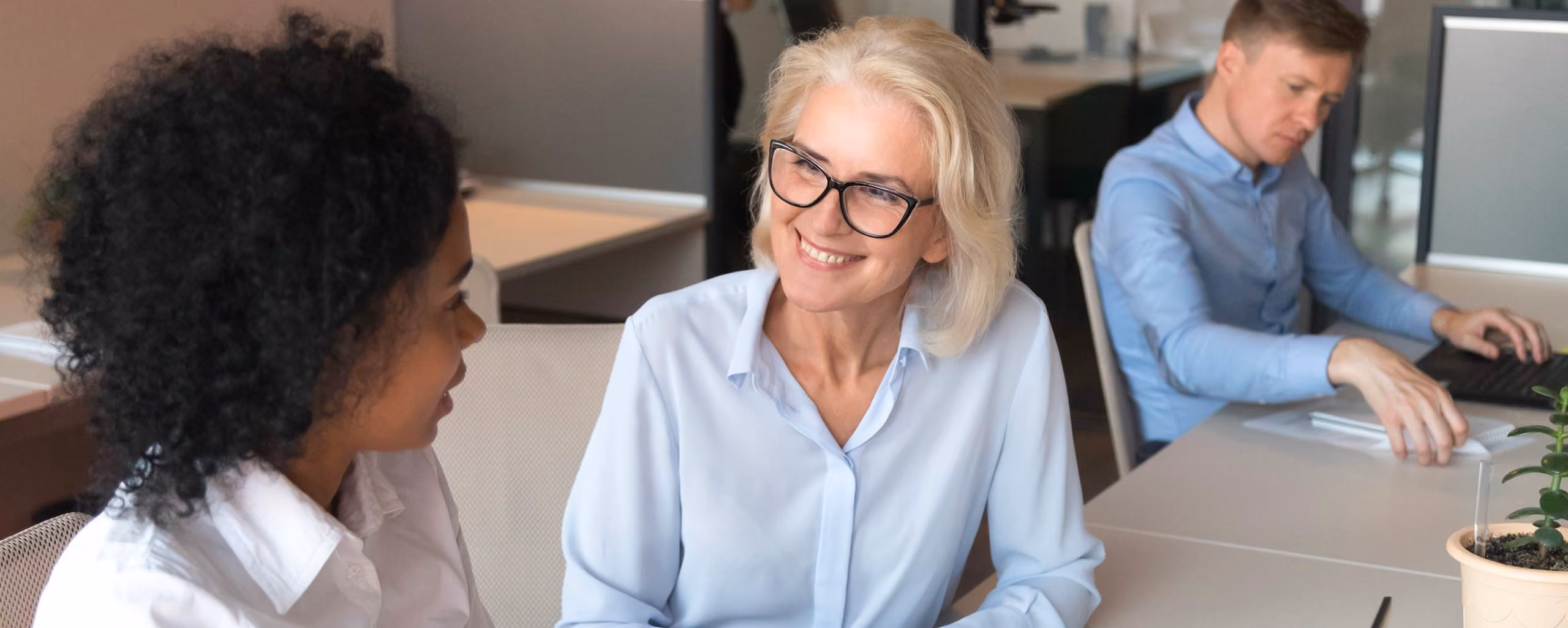 Two women smiling and having a conversation in an office, with a man working on a laptop nearby.