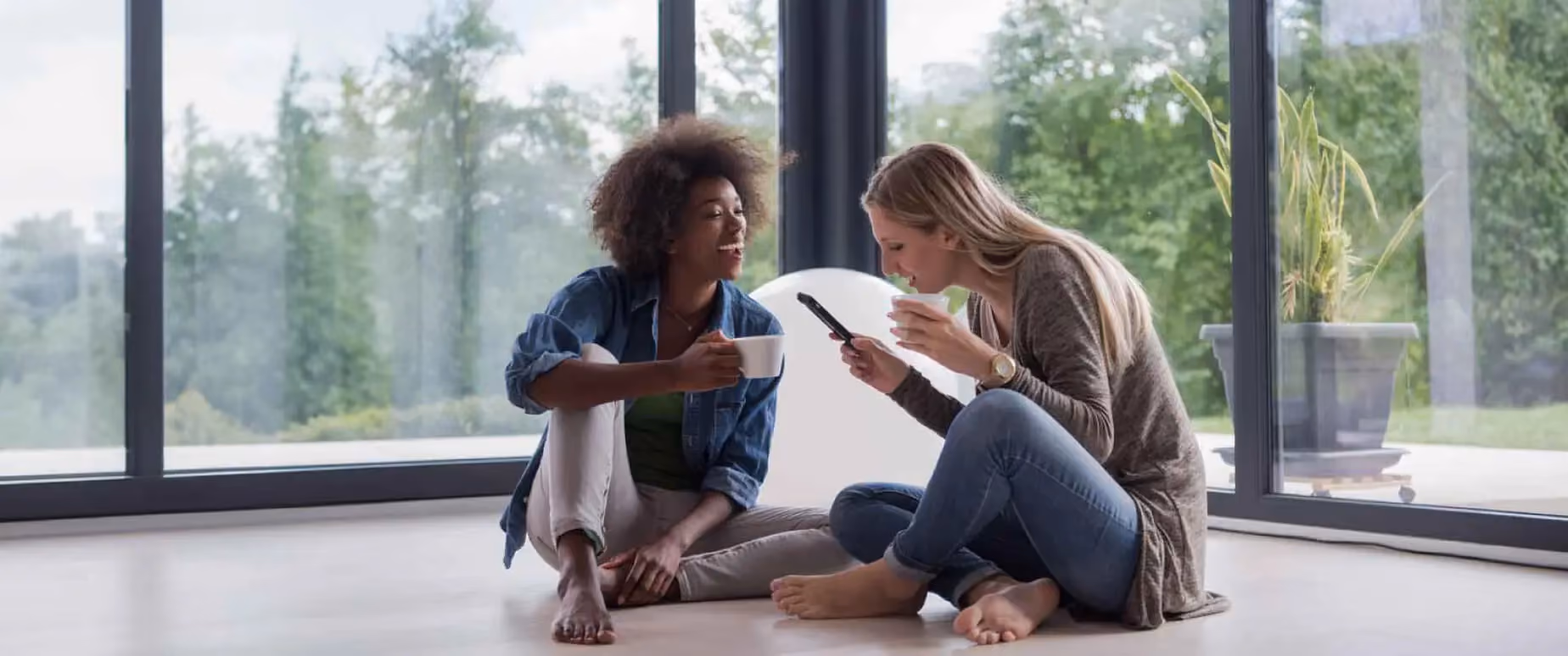 Two women sit on the floor of a living room while expressing joy over something they are reading on their phone.