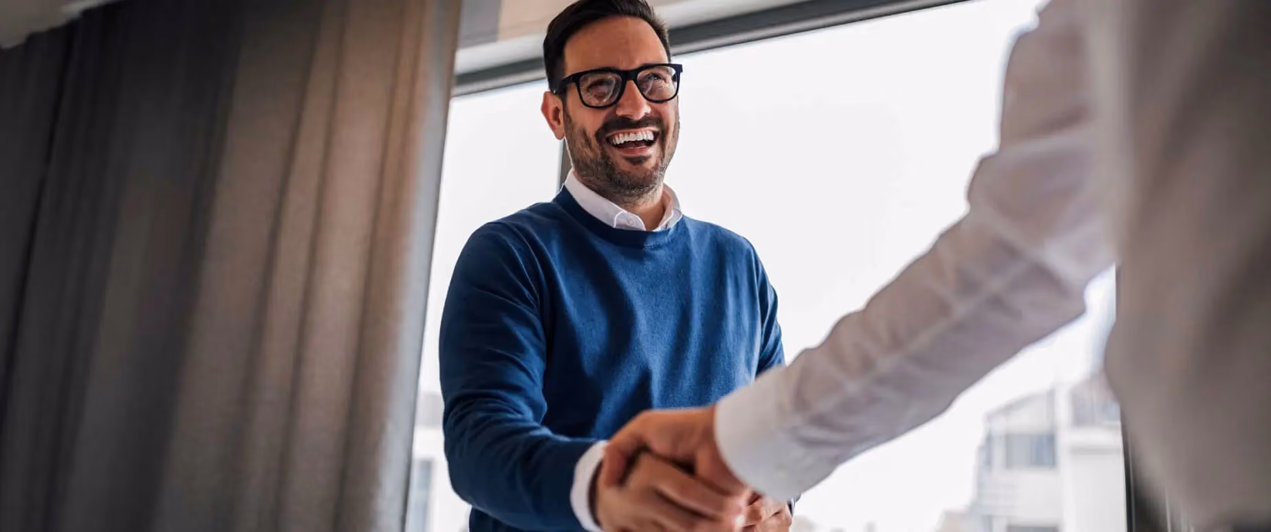 A man wearing a blue sweater and black glasses is smiling and shaking the hands of his rental client.