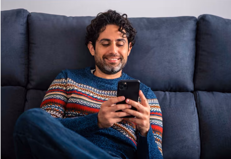 Man in patterned sweater sitting on sofa, smiling while using a smartphone.