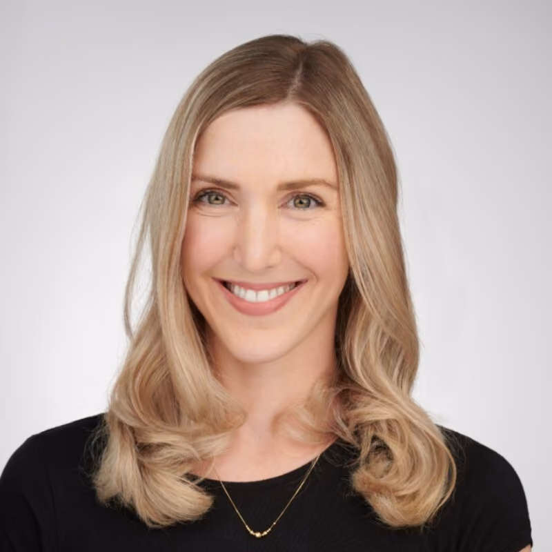 Smiling woman with blonde wavy hair wearing a black top and a gold necklace on a gray background.