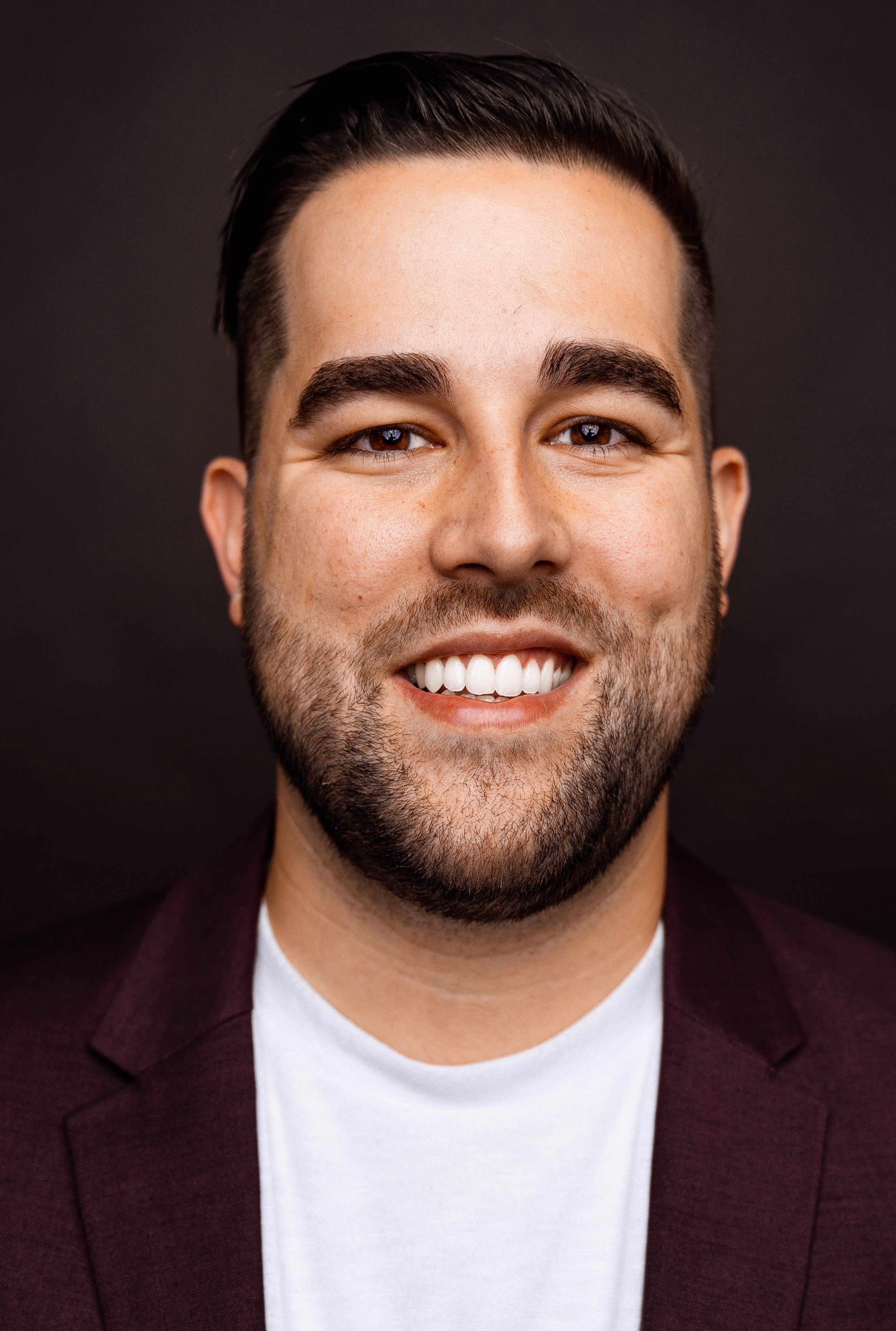 Smiling man with dark hair, beard, and maroon blazer on a dark background.