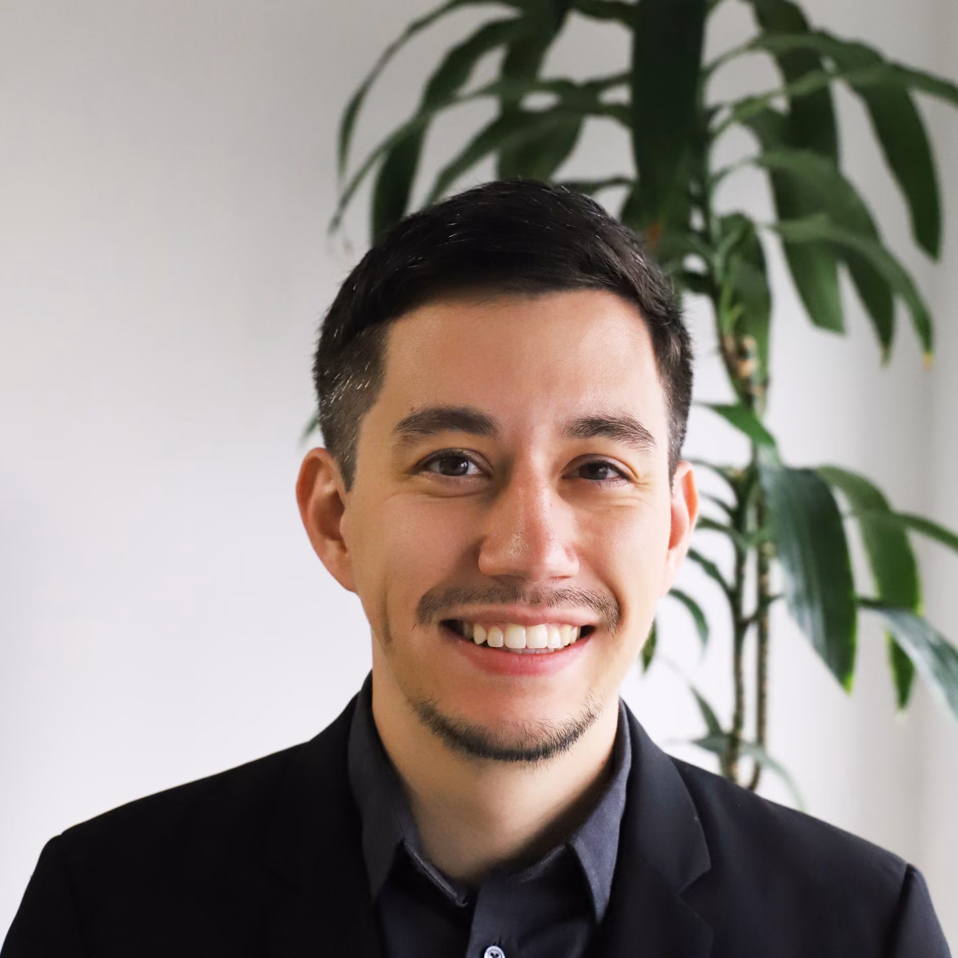 Smiling man with short dark hair and a trimmed beard wearing a black blazer with a plant in the background.