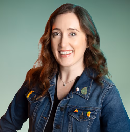 Smiling woman with wavy brown hair wearing a denim jacket with colorful pins against green background.