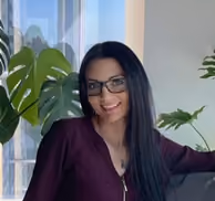 Woman with long black hair and glasses smiling, seated near window with large green plants.