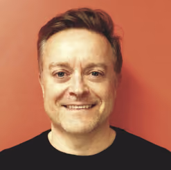 Smiling man with short brown hair and light stubble wearing a black shirt against a red background.