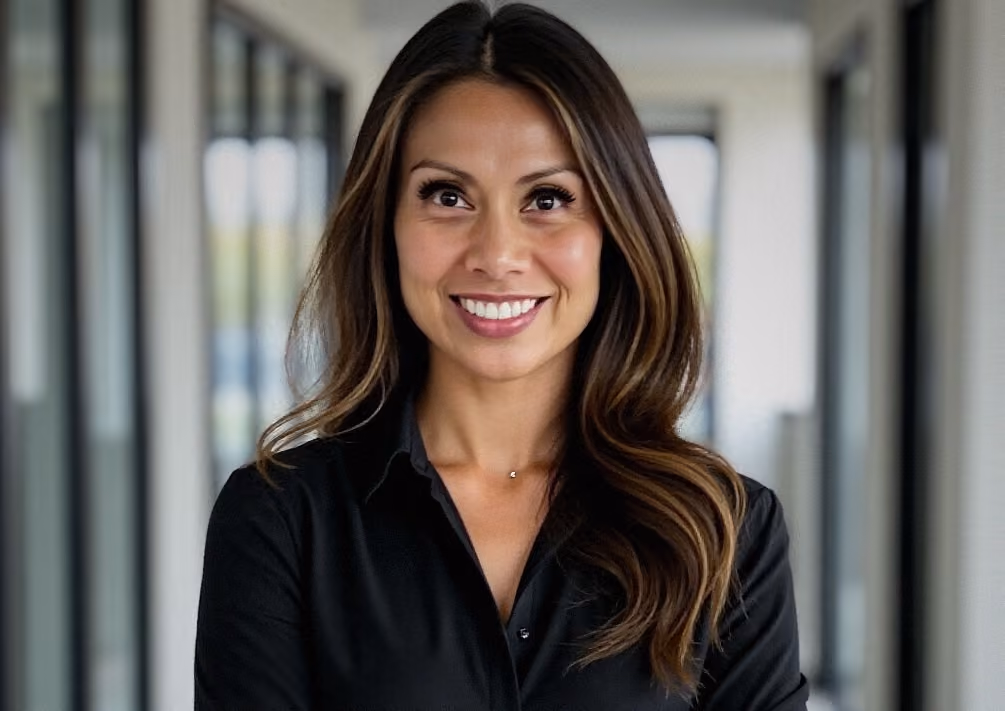 Smiling woman with long brown hair in a black shirt standing in a bright modern hallway.