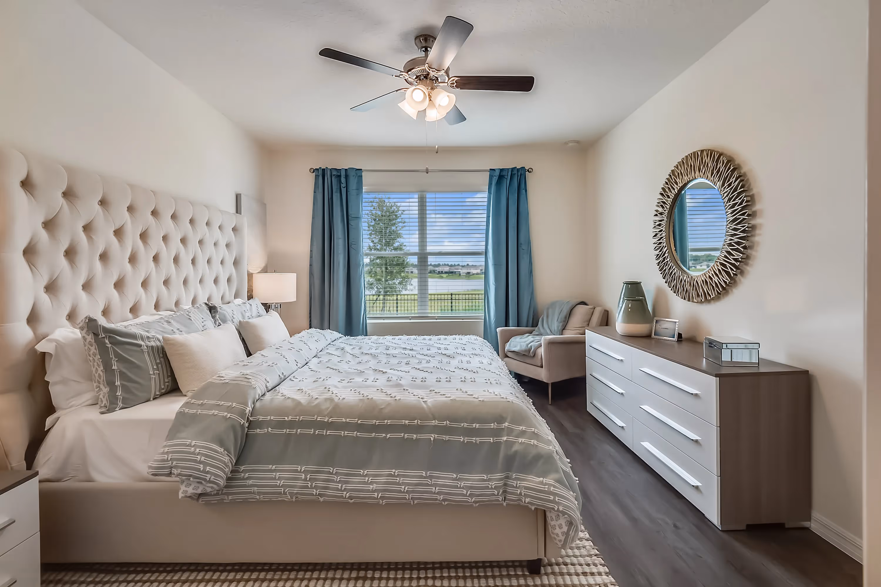 Elegant master bedroom in a Landing at Stone Chimney home, Supply NC, with a tufted headboard, blue curtains, a large window, and a modern dresser