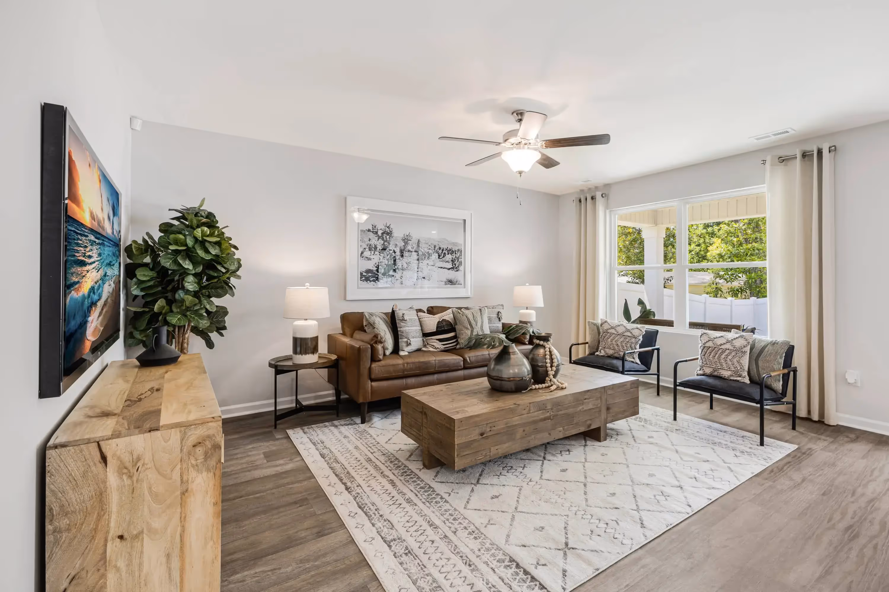 Cozy living room featuring a brown sofa with decorative pillows, a wooden coffee table, and black metal chairs. Light-colored walls and large windows provide natural light. A ceiling fan hangs above, and a stylish area rug lies on the floor. Decorative items include a plant, lamps, and artwork on the wall.