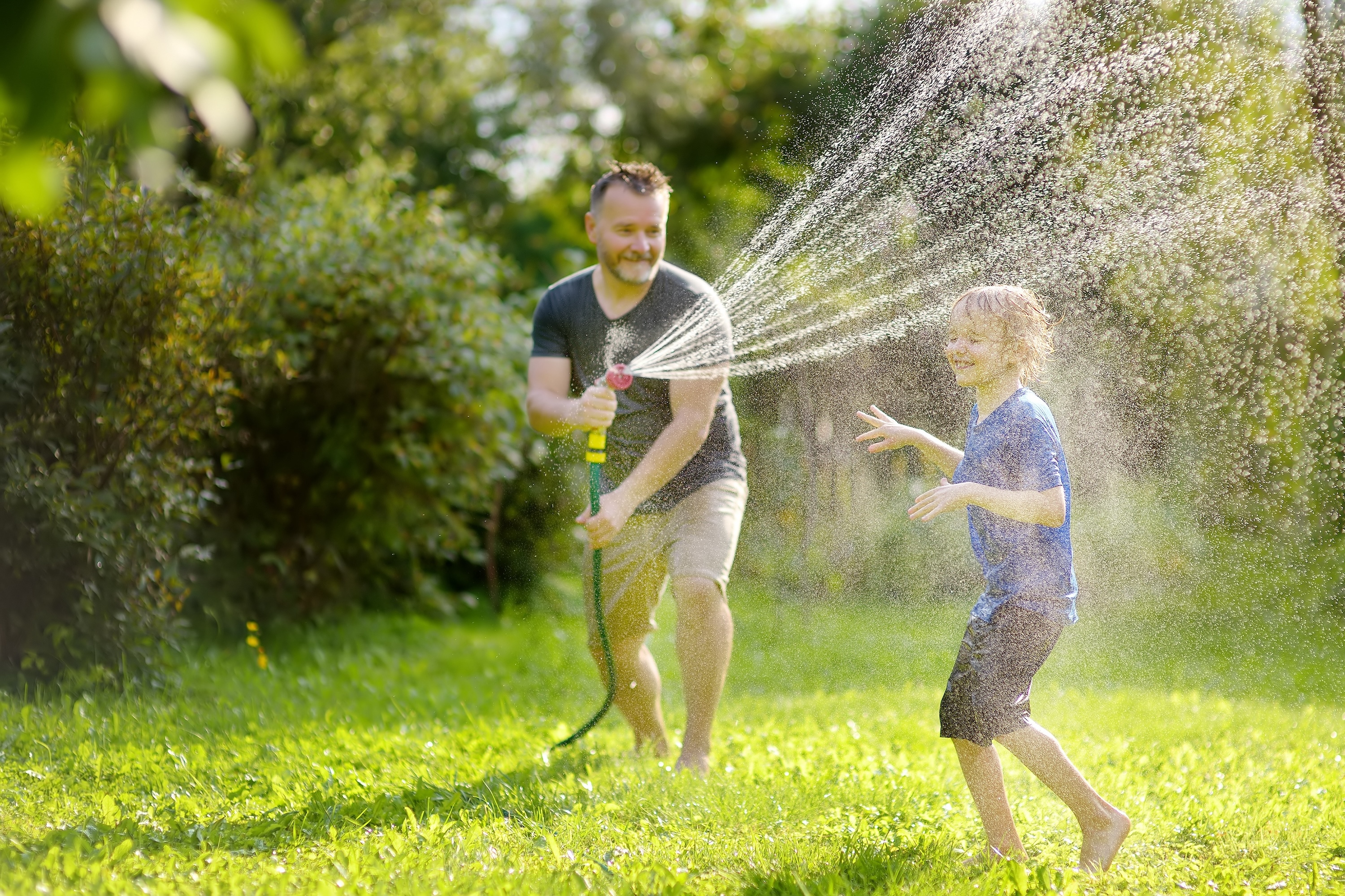 Smiling father spraying water on his happy son with a garden hose in a green backyard, depicting family fun at Landing at Stone Chimney homes in Supply, NC