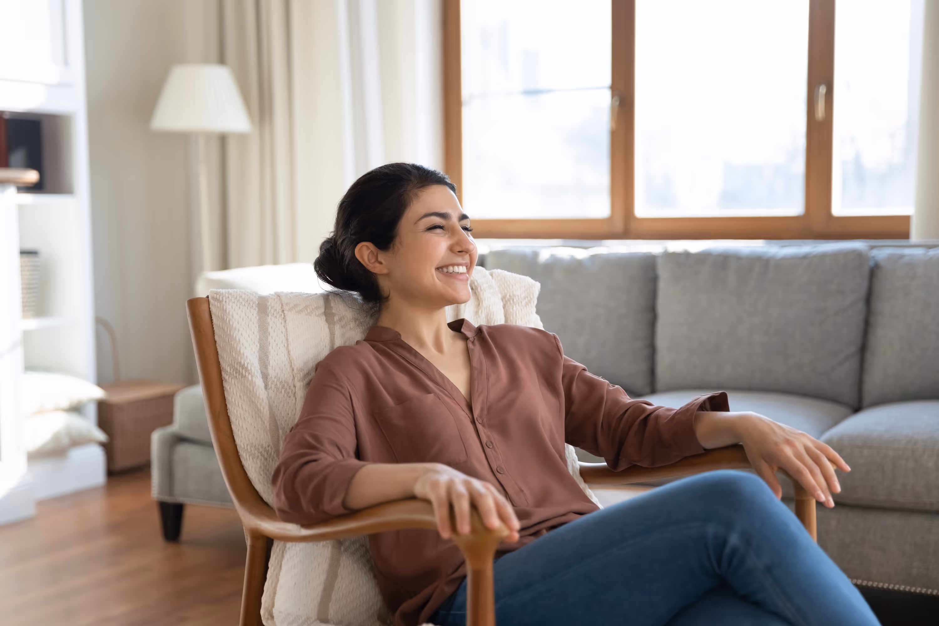 A smiling woman relaxing in a comfortable chair in a bright, sunlit living room, depicting serene home life at Landing at Stone Chimney in Supply, NC.