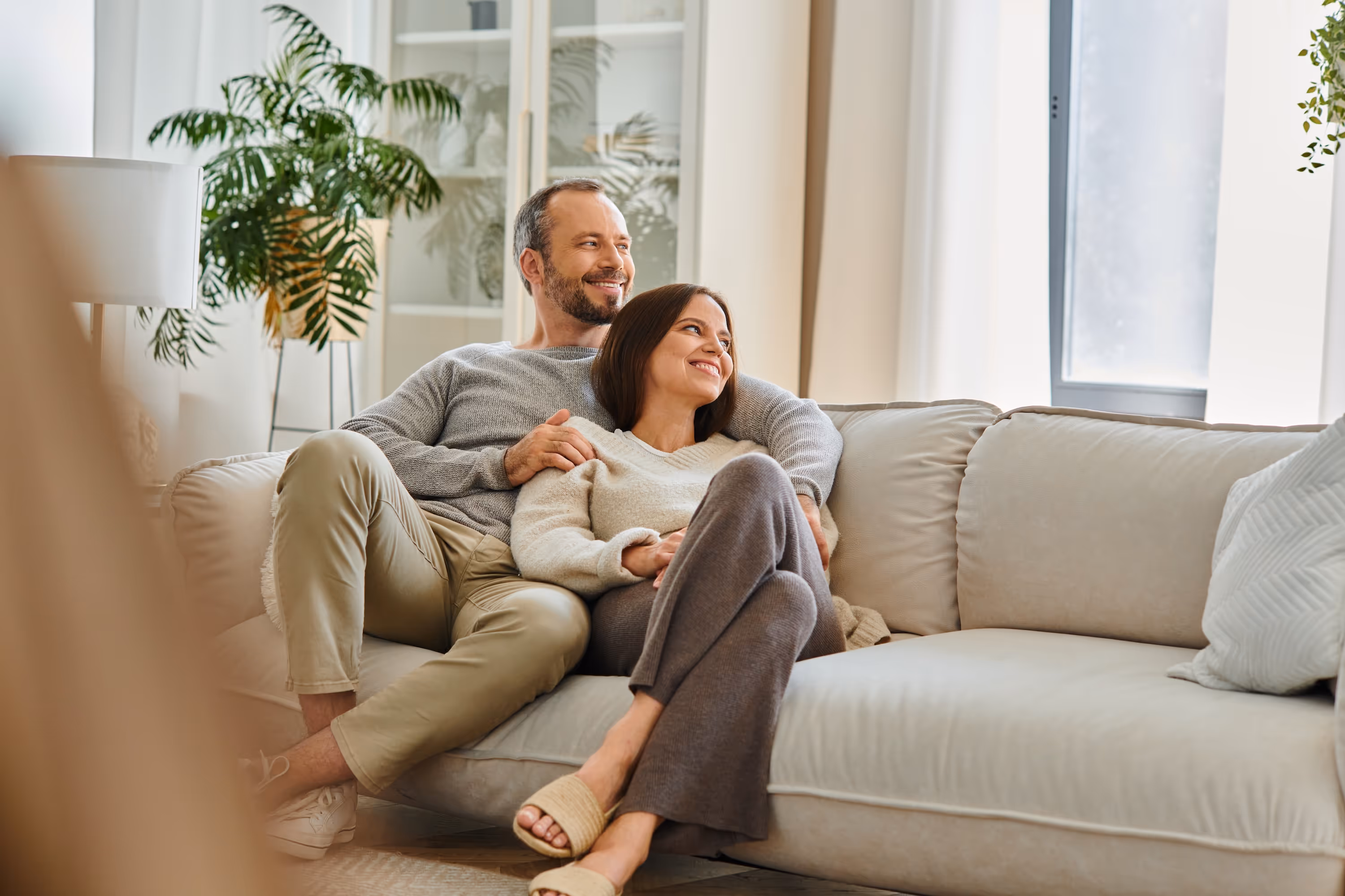 A happy couple smiling and relaxing on a comfortable sofa in a sunlit living room, representing a serene home life at Landing at Stone Chimney in Supply, NC.