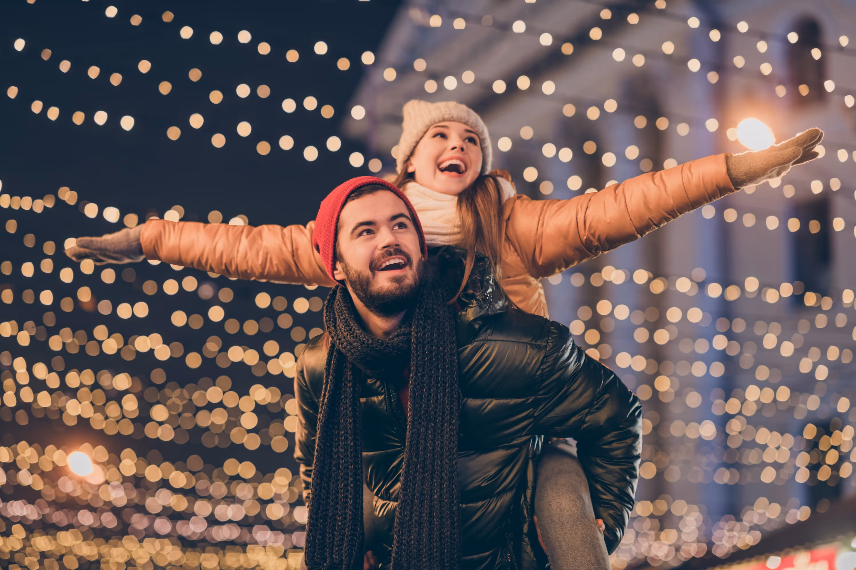 A smiling couple dressed in warm winter clothing enjoying an evening outdoors near The Landing at Stone Chimney, surrounded by glowing holiday string lights.