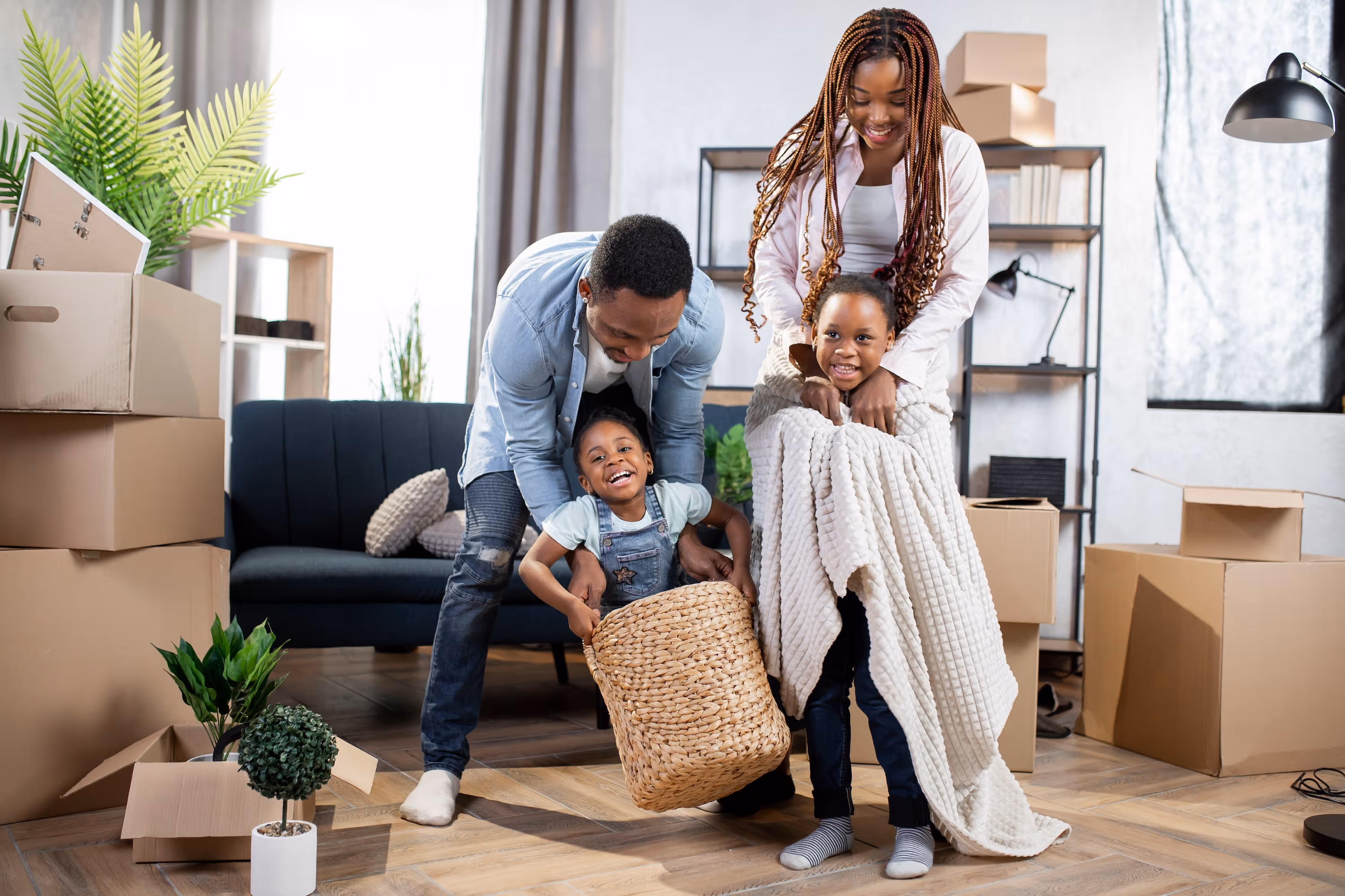 Two adults playing with children using a laundry basket and blankets in a room with moving boxes at The Landing at Stone Chimney.
