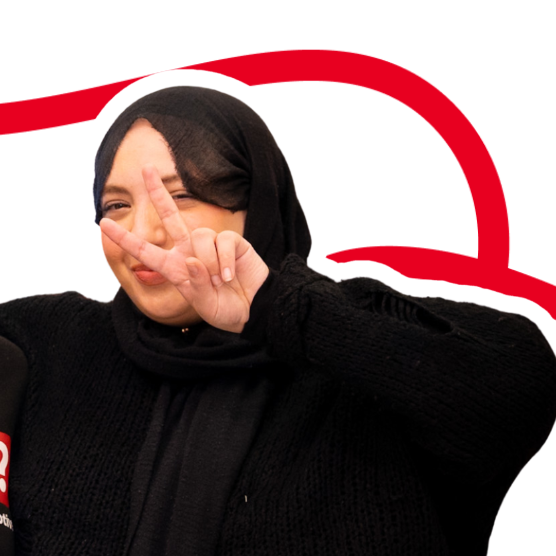 Woman wearing a black hijab and sweater making a peace sign with her right hand while smiling.