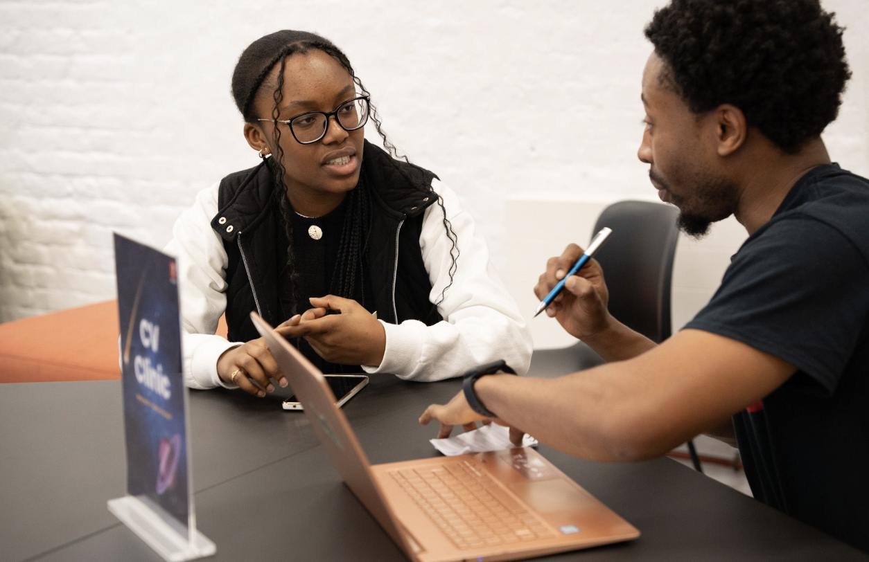 Two people seated at a table discussing, with a laptop and a CV clinic sign on the table.