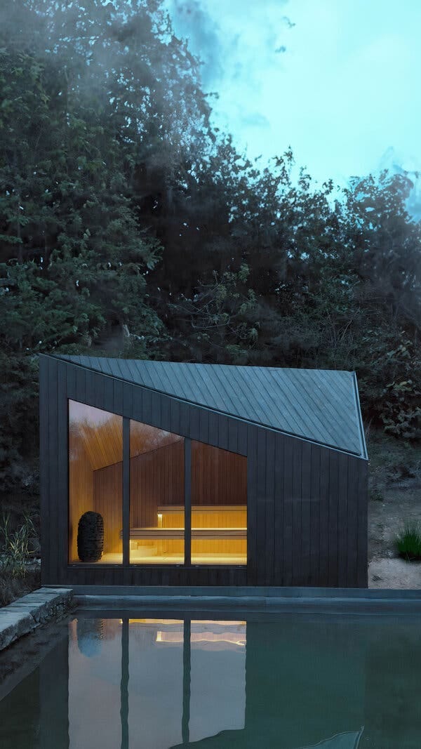 A cabin with a triangular roofline and a trio of windows glows from within. In front of it is a pool, behind it, trees.