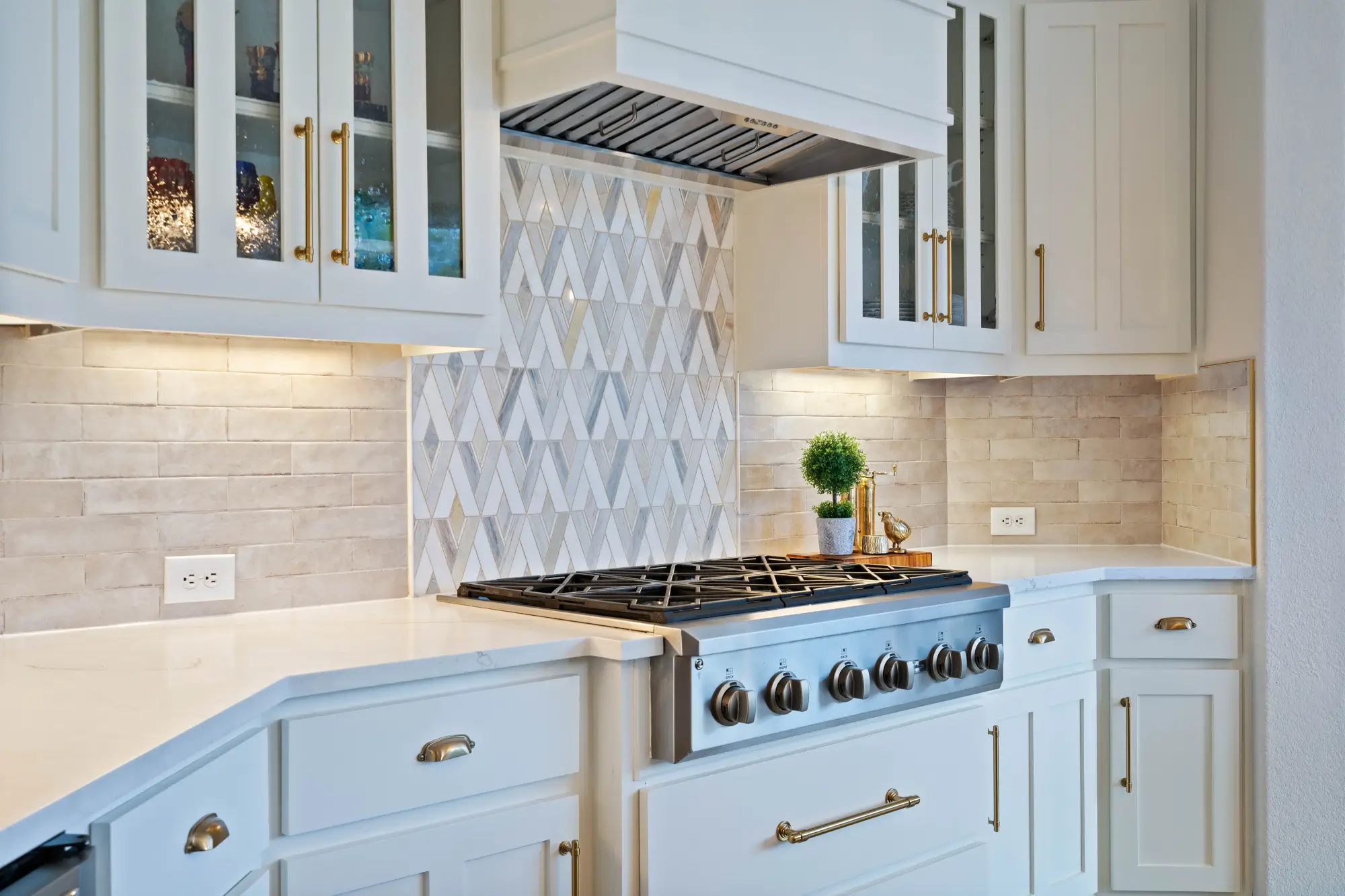 Modern kitchen with white cabinetry, gold hardware, a stainless steel gas stove, and a geometric tile backsplash. Under-cabinet lighting illuminates the countertops, and a small potted plant and utensils sit beside the stove.