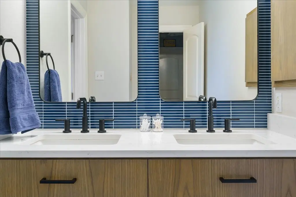 Bathroom with double vanity, white countertop, wood cabinets, black faucets, and two mirrors mounted on a blue tiled backsplash. Blue towels hang on black towel rings on each side.
