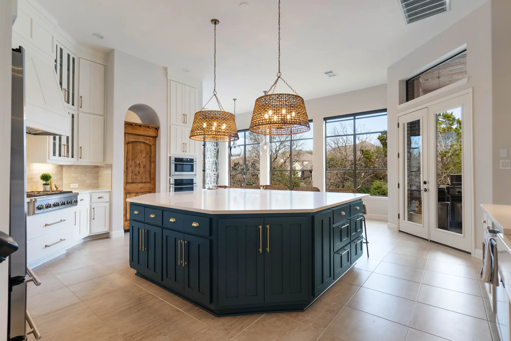 Spacious kitchen with large navy island, gold hardware, white countertops, wicker pendant lights, white cabinetry, and large windows overlooking a backyard. A wooden door and outdoor patio area are visible.