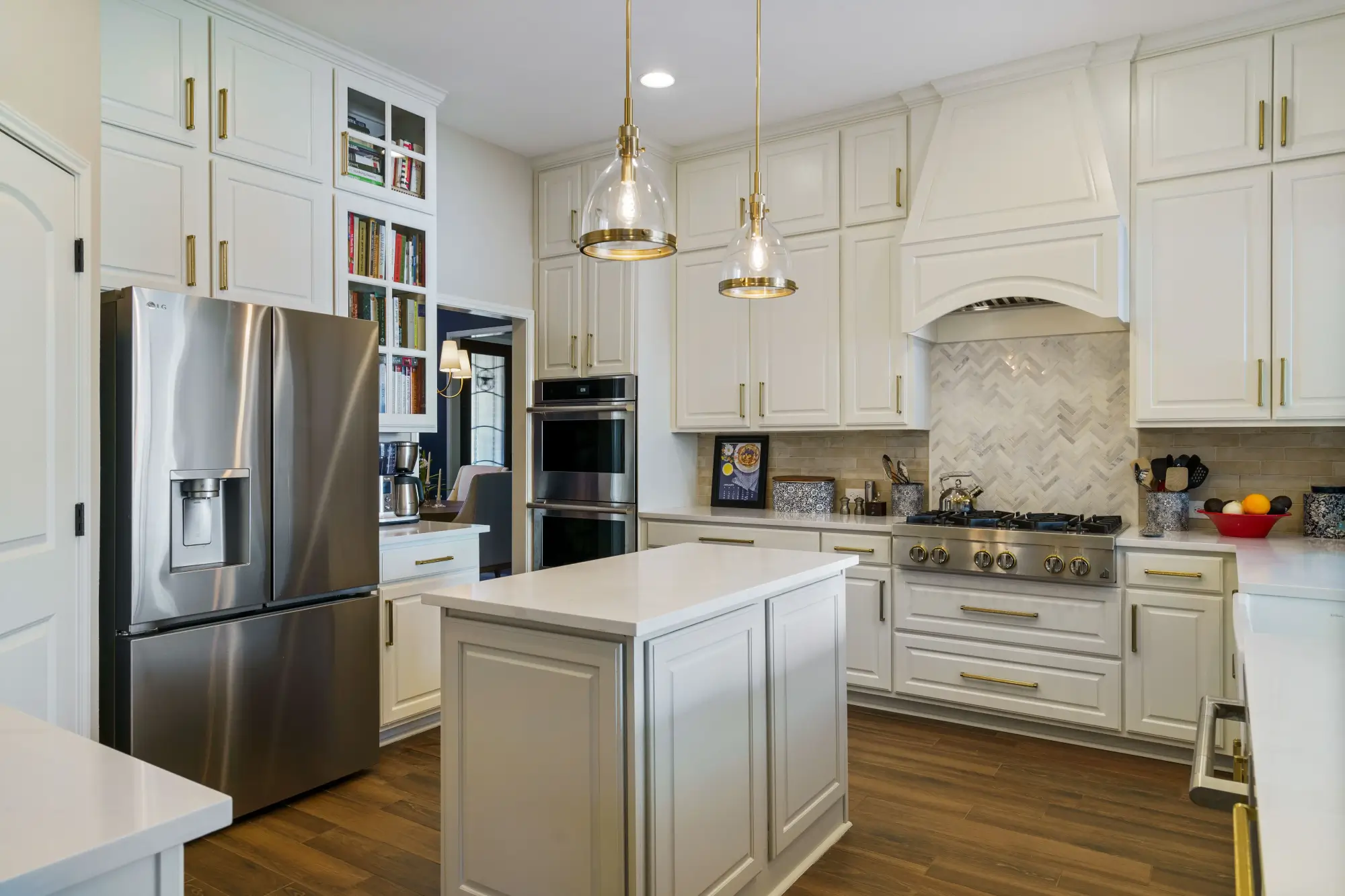 Elegant kitchen with white cabinetry, gold hardware, stainless steel appliances, small island, glass pendant lights, and wood flooring. Herringbone tile backsplash behind the stove and built-in bookshelves with cookbooks.