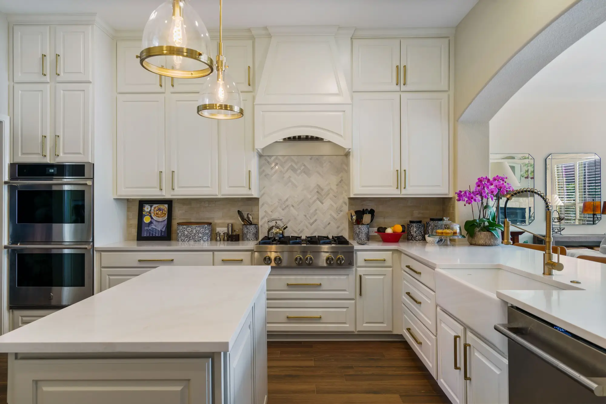Modern kitchen with white cabinetry, gold hardware, a large island, glass pendant lights, wood flooring, and a farmhouse sink. Stainless steel double oven and gas stove with herringbone tile backsplash. Purple orchid on the counter.