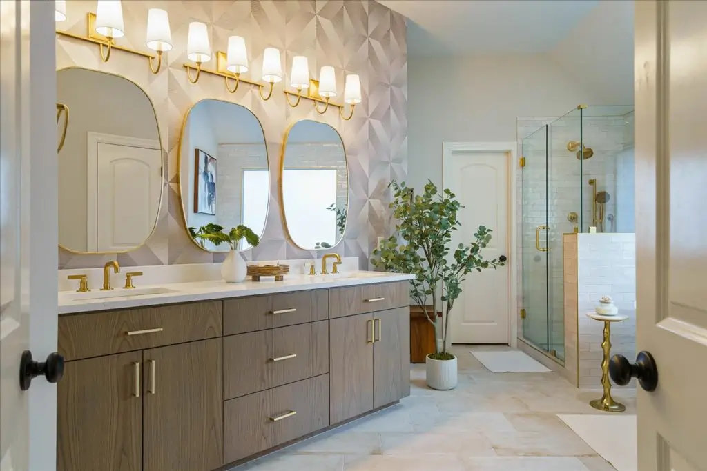 Elegant bathroom with double vanity, wood cabinetry, gold fixtures, and three arched mirrors. A geometric accent wall, glass shower enclosure, white tiles, and potted plant add modern touches. Sconces above the mirrors provide lighting.