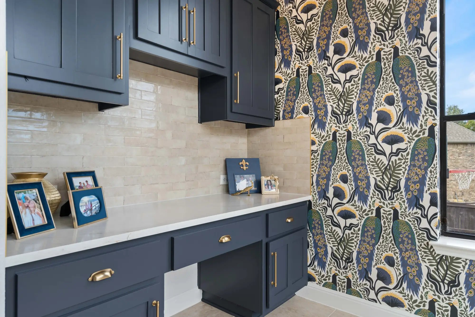 Home office nook with navy cabinetry, gold hardware, white countertop, beige tile backsplash, and bold peacock-patterned wallpaper. Framed photos and gold decor sit on the counter, with a window providing natural light.
