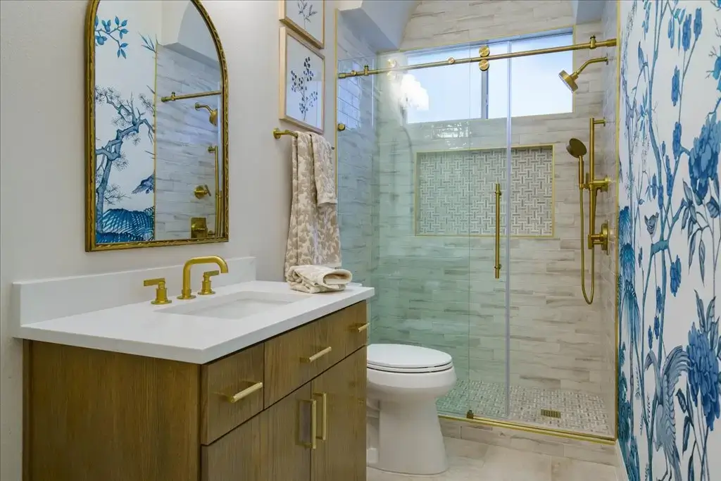 Elegant bathroom with wood vanity, gold hardware, white countertop, arched mirror, blue floral wallpaper, and glass shower with gold fixtures. Beige tile walls and a decorative niche accent the shower.