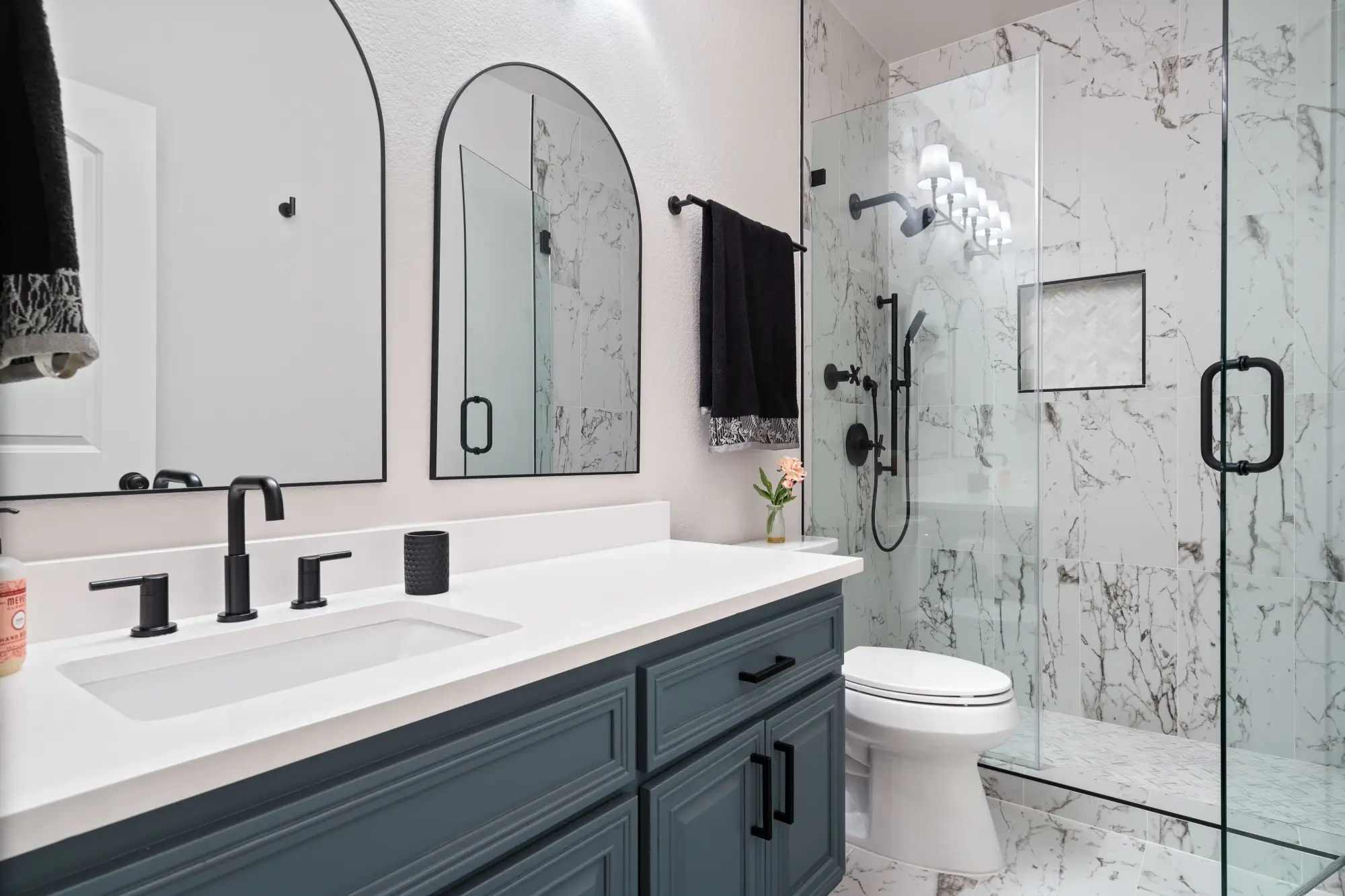 Modern bathroom with navy vanity, white countertop, black fixtures, arched mirrors, and a glass shower with marble tile walls. Black towel hangs beside the shower, and a small flower vase adds decor.