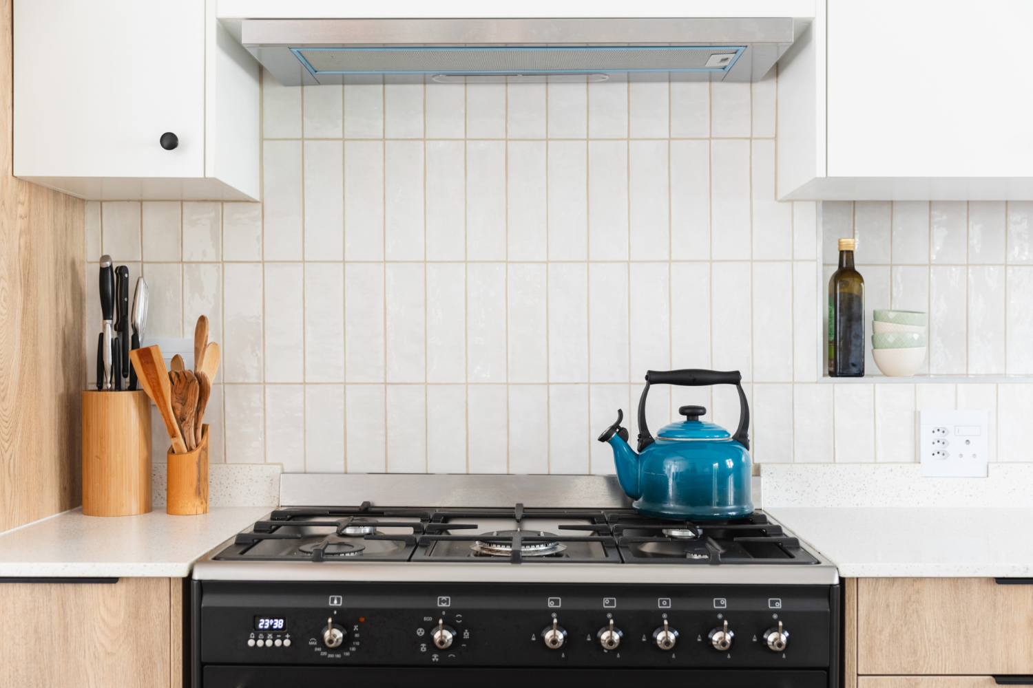 Modern kitchen with a gas stove, a blue kettle on the burner, a range hood above, and light tiled backsplash. To the left are wooden utensils and knives in holders, and to the right are a bottle of olive oil and stacked bowls on a small shelf.