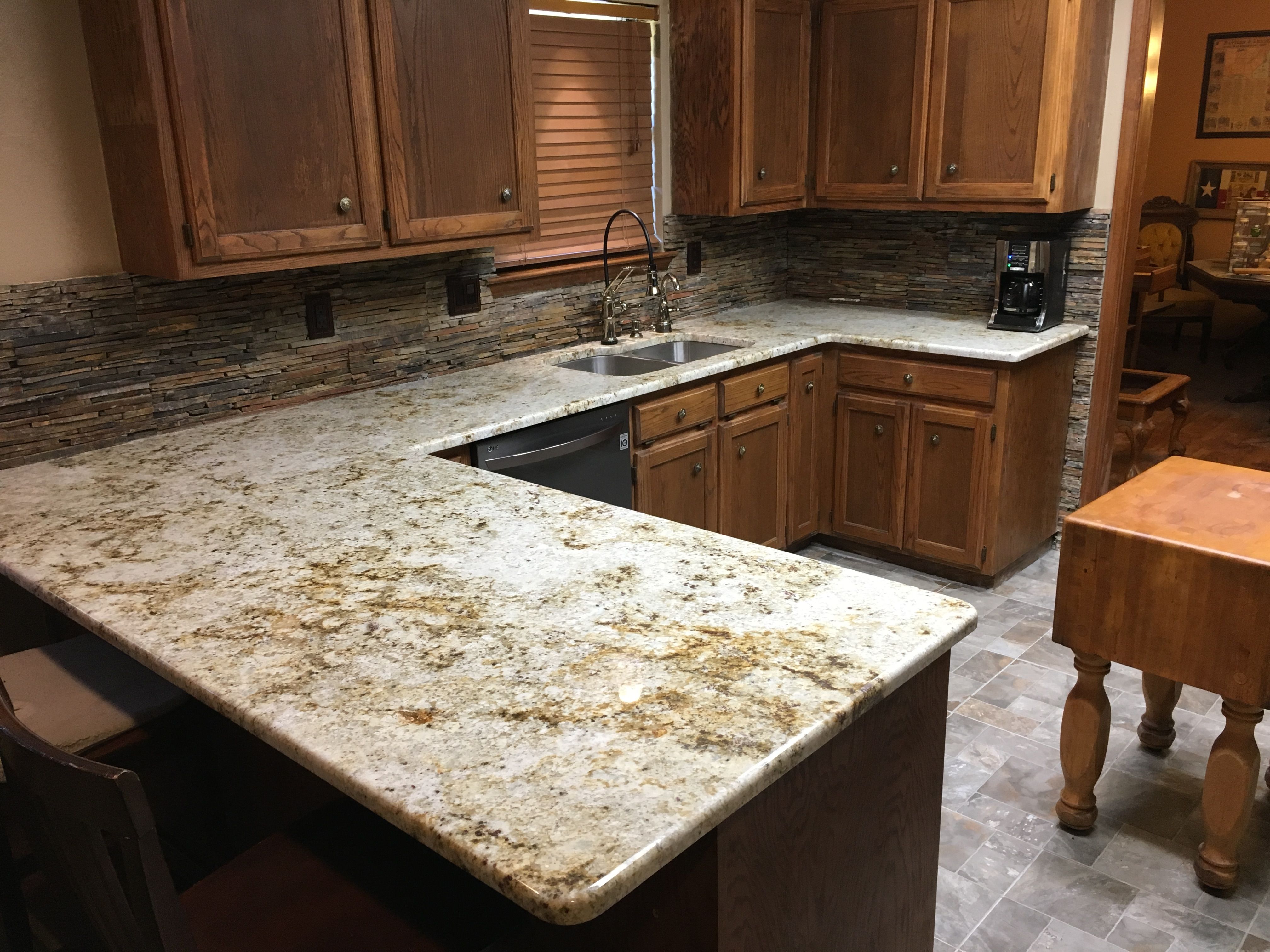 Traditional kitchen with wooden cabinets, speckled granite countertops, and stacked stone backsplash. A stainless steel sink with a gooseneck faucet is under a window with wooden blinds. There is a coffee maker on the counter and a small butcher block isla
