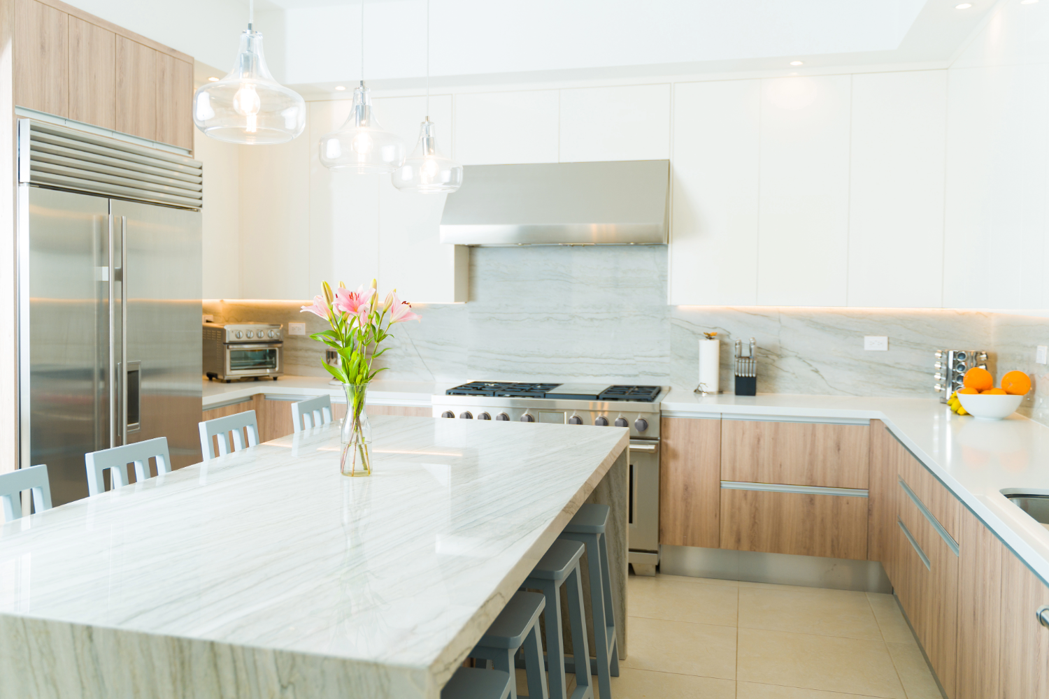 Bright modern kitchen with a large marble island, gray chairs, and a vase of pink lilies. Stainless steel appliances include a fridge, oven, and range hood. Light wood and white cabinetry line the walls, and countertops hold a knife block, toaster oven, pa