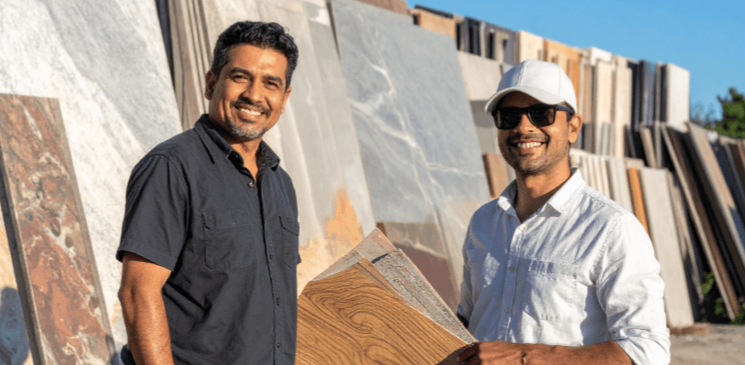 Two men standing in front of large stone slabs, one holding samples of stone and wood, both smiling.