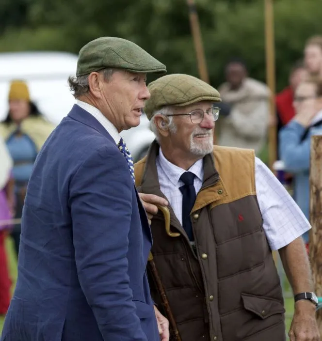 Two older men wearing flat caps and outdoor clothing, one in a blue blazer and patterned tie, engaged in conversation outdoors during an event.