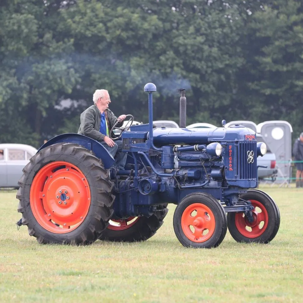 A man sitting in an old, blue tractor with red wheels driving in an exhibition.