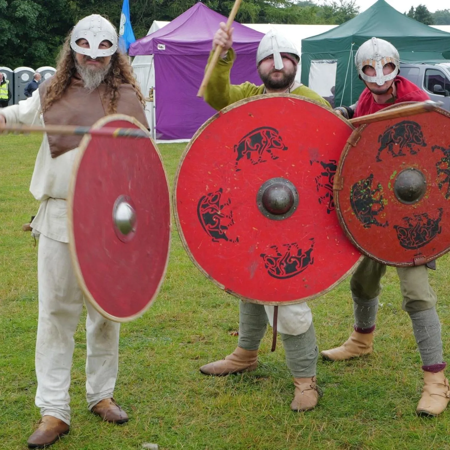 Three men in Viking helmets and medieval attire holding round red shields with black designs at an outdoor event.