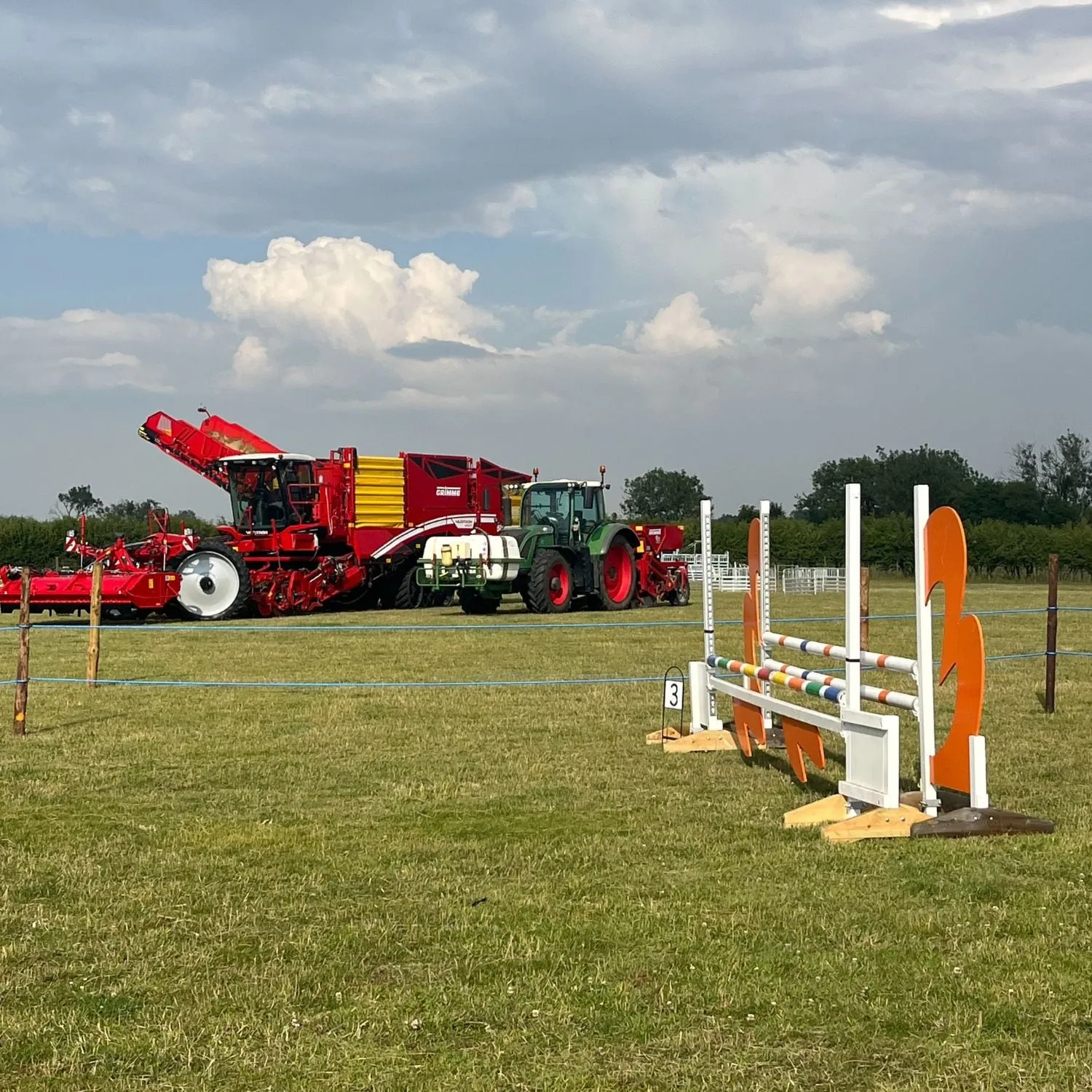 Red and yellow agricultural harvester machine and tractor on a grassy field under a partly cloudy sky near an equestrian show jumping obstacle.