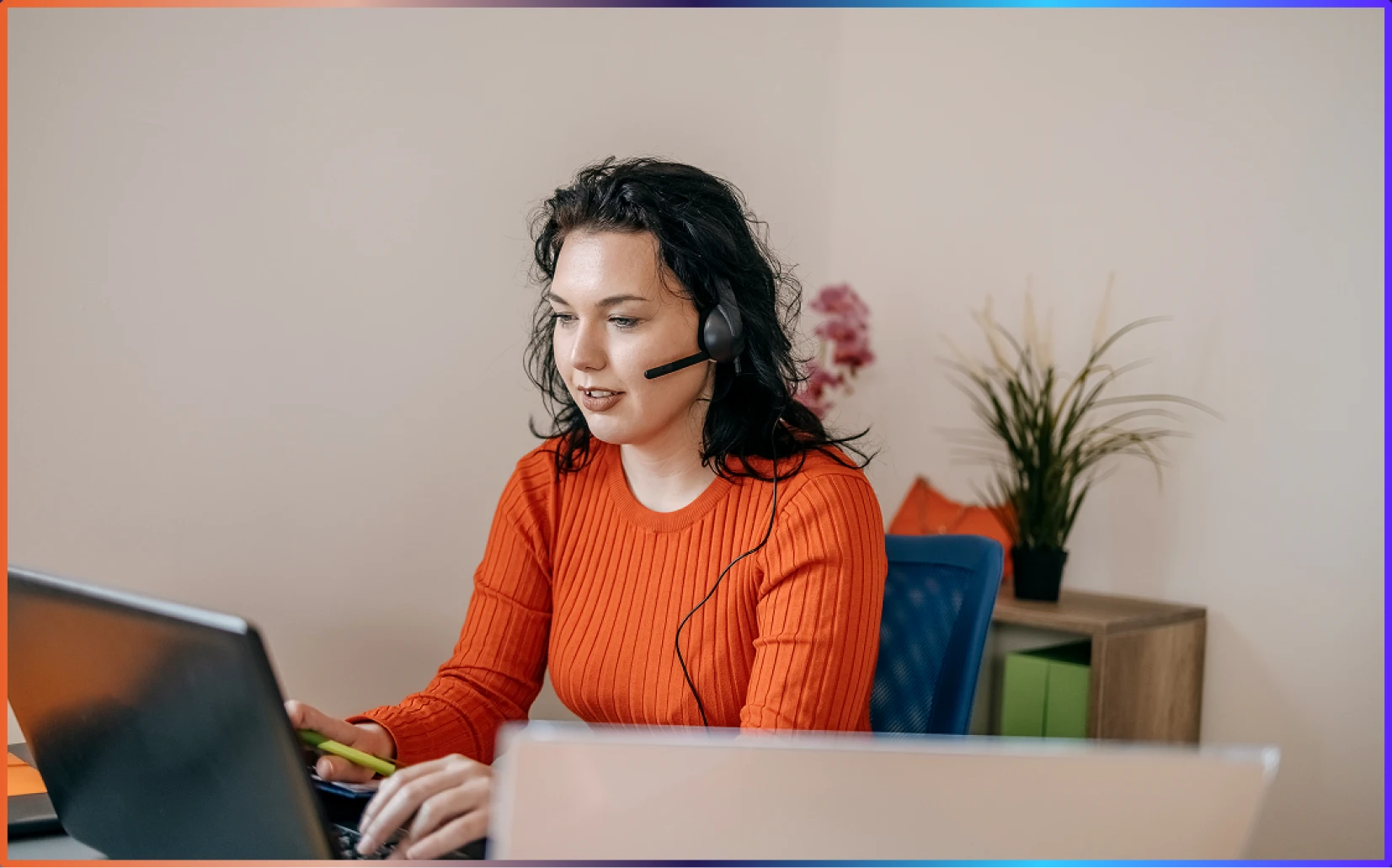 Woman with headset in an orange sweater working on a laptop in an office setting.