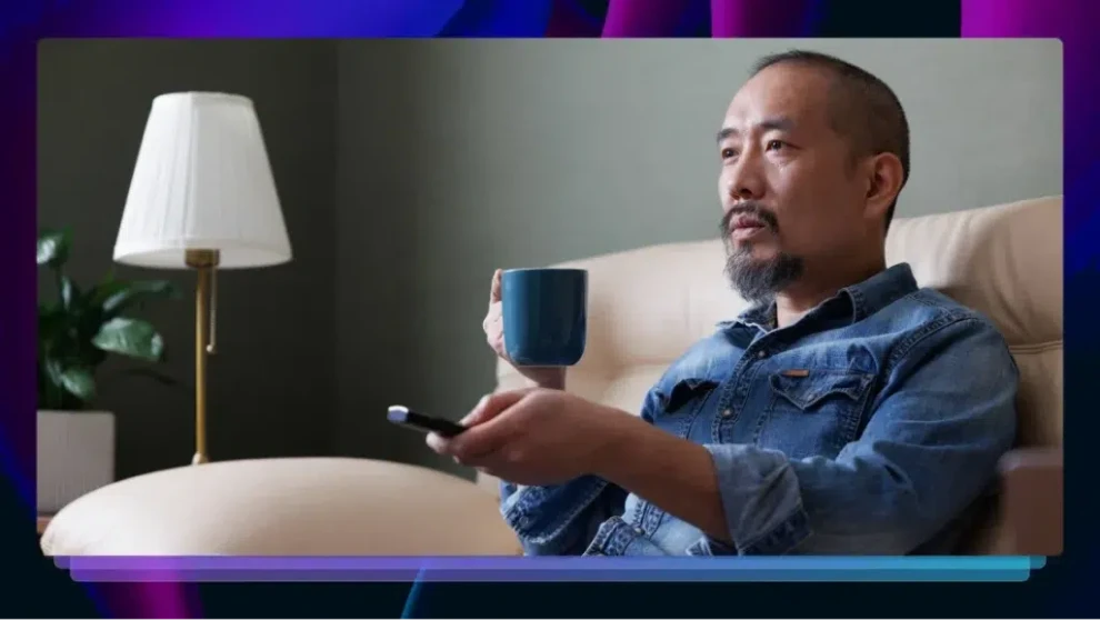Man watching television while holding a remote and coffee, representing viewing video content at home.