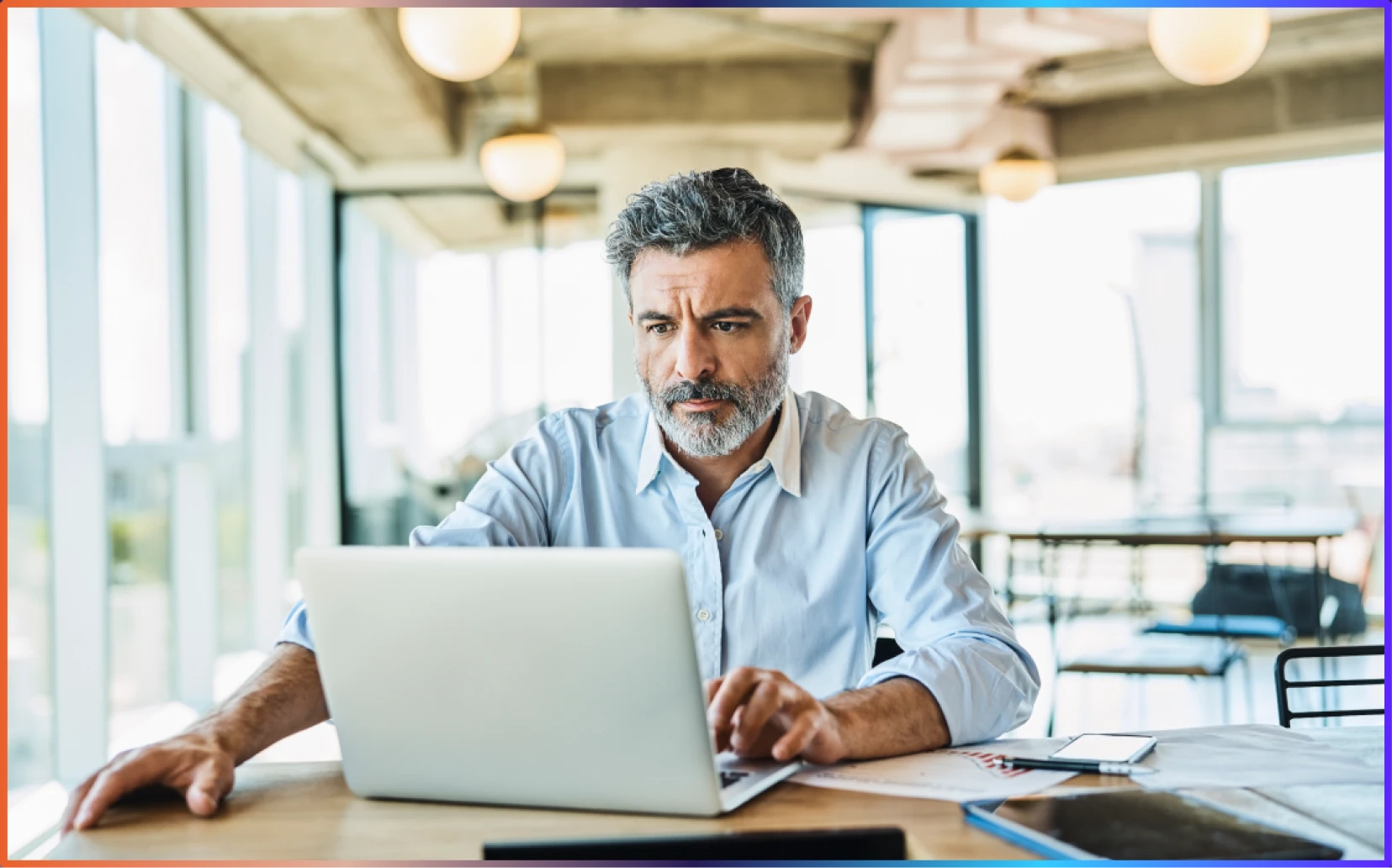 Man with gray hair and beard focused on working on a laptop at a bright modern office desk.