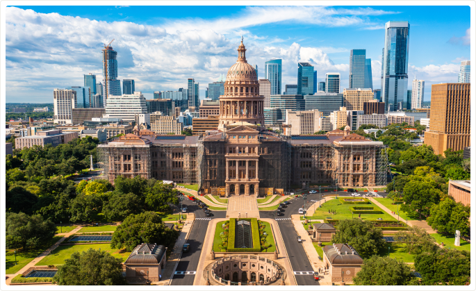 Aerial view of the Texas State Capitol building with Austin skyline in the background on a partly cloudy day.