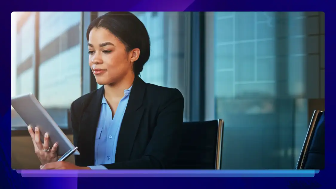 Young woman in a blazer holding and looking at a tablet in a modern office setting.