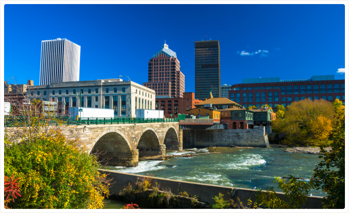 City skyline with tall buildings and an arched stone bridge over a flowing river on a clear day.