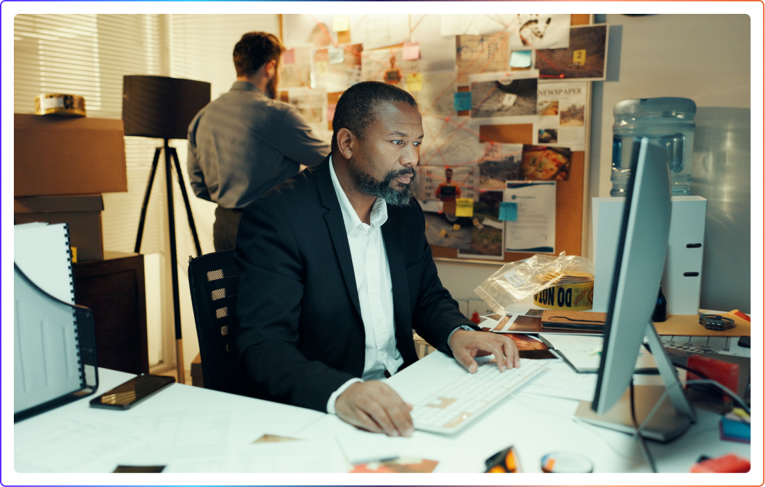 Professional at a desk working at a computer, with an evidence board of pinned photos and documents on the wall behind him.