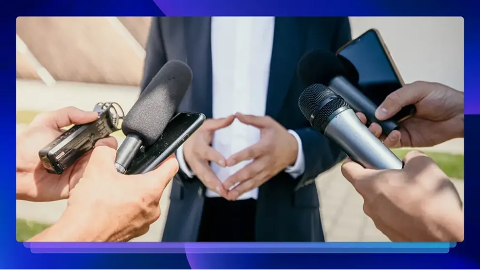 Person in suit being interviewed with microphones and smartphones held up around them.