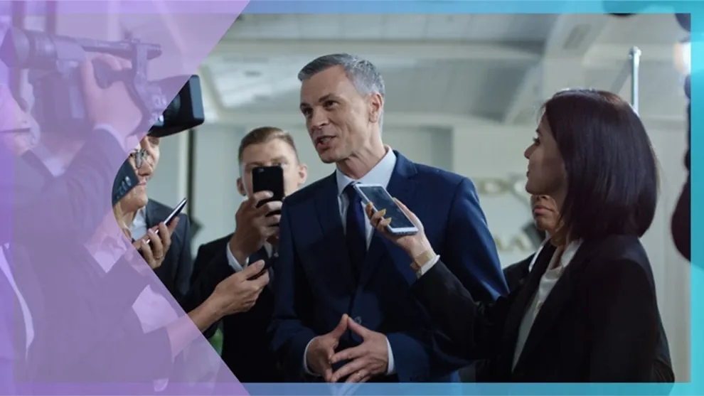 Man in suit speaking to reporters holding phones and a microphone in an indoor setting.