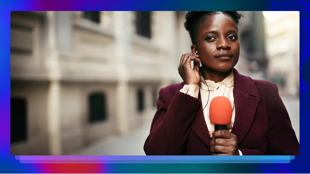 Female reporter adjusting earpiece, holding orange microphone, standing on city street near a building.